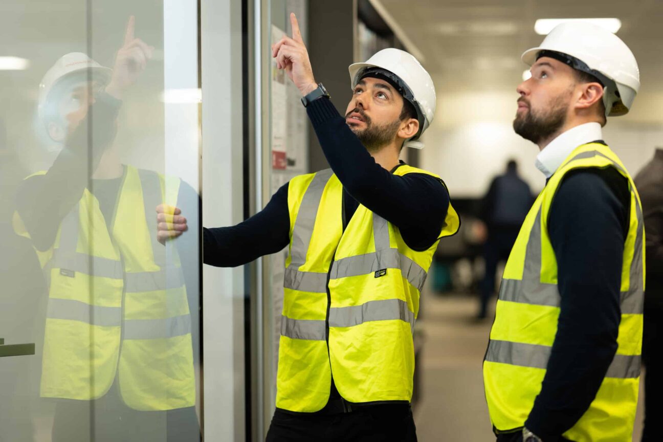 Two men wearing hi-vis vests and white hard hats, pointing at a glass door panel